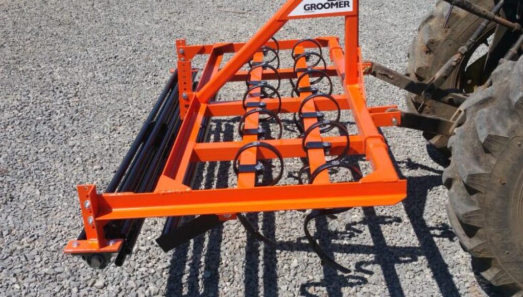Orange arena groomer attachment showing spring tines and leveling rollers on gravel surface